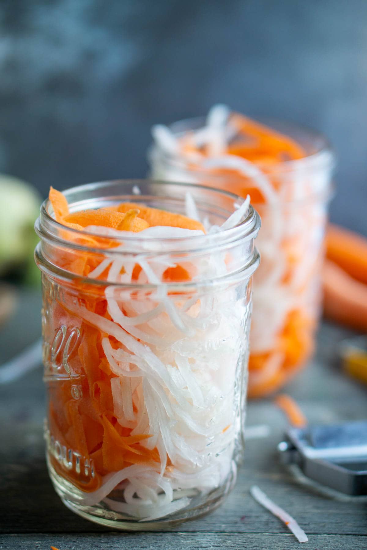 Two mason jars filled with shredded carrots and daikon radish sit on a wooden surface with a peeler nearby.