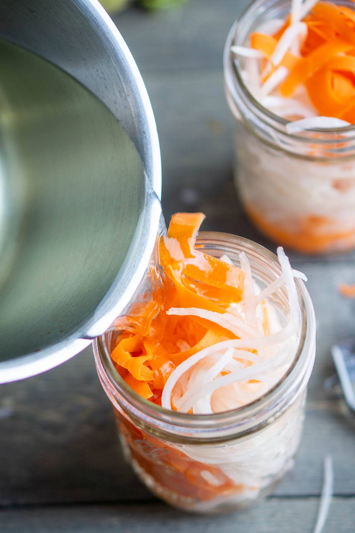 A metal pot pours liquid into a glass jar filled with sliced carrots and daikon radish on a wooden surface.