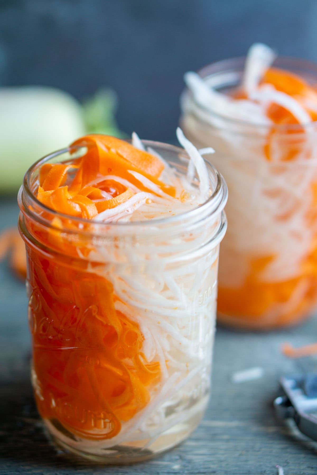 Two glass jars filled with spiralized carrots and daikon radish in a pickling liquid, placed on a wooden surface.
