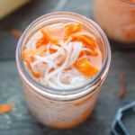 A jar filled with pickled vegetables, including thinly sliced white radish and orange carrot strips, in a light brine.