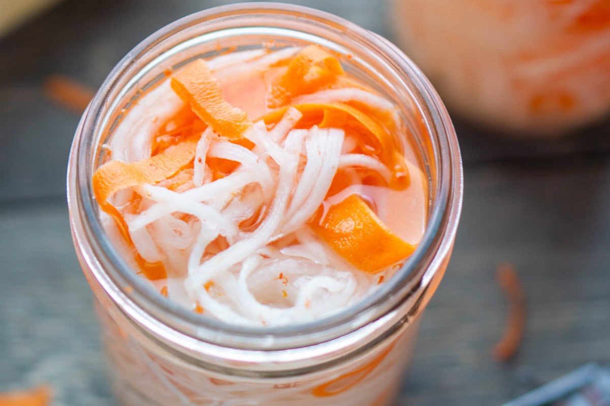 A jar filled with pickled vegetables, including thinly sliced white radish and orange carrot strips, in a light brine.