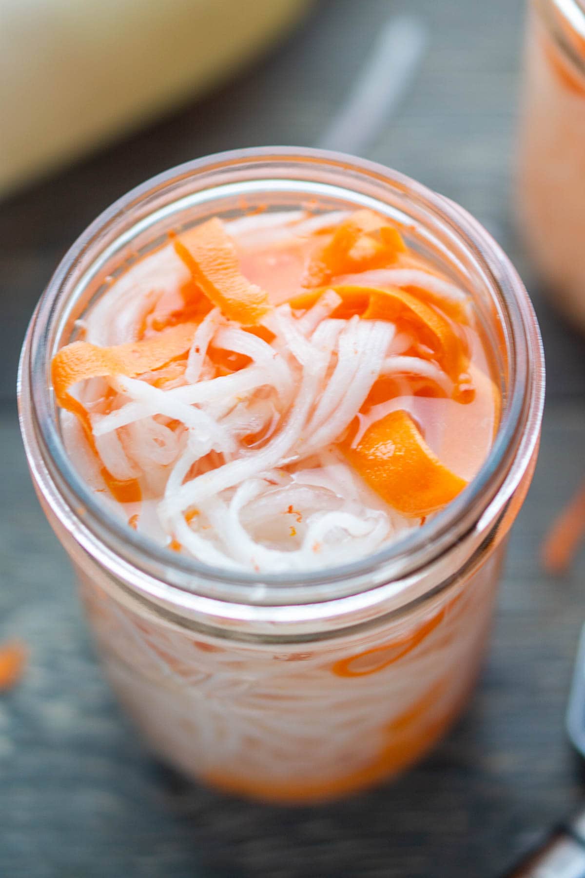 A jar filled with pickled vegetables, including thinly sliced white radish and orange carrot strips, in a light brine.