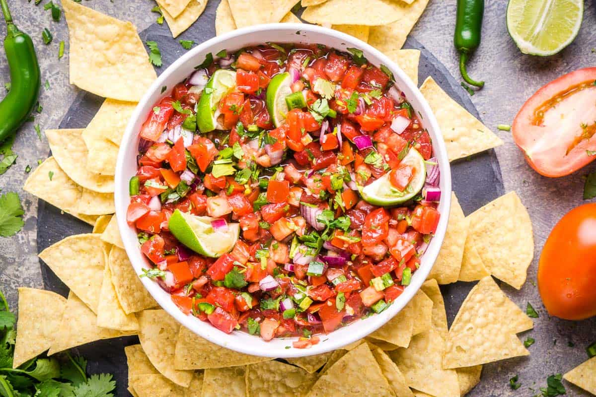 pico de gallo in white serving bowl surrounded by tortilla chips, tomatoes, lime wedges, and serrano peppers
