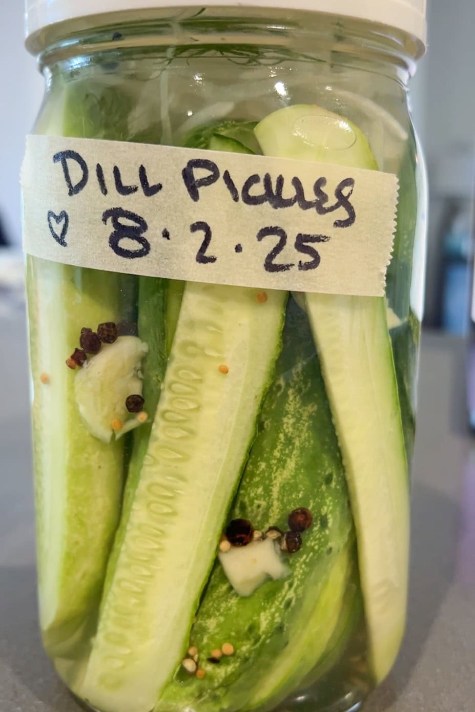 A glass jar filled with sliced cucumbers, spices, and garlic, labeled Dill Pickles 8.2.25 on a piece of tape.