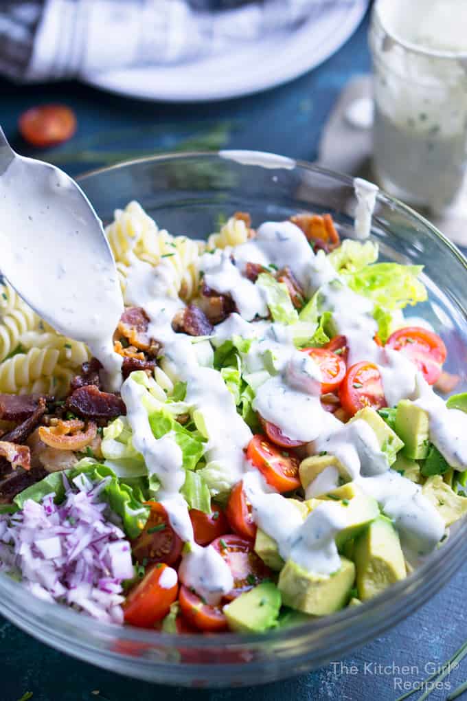 blt pasta salad with spoon drizzling ranch dressing in glass bowl on blue surface with plates, napkin, and ranch dressing in a jar