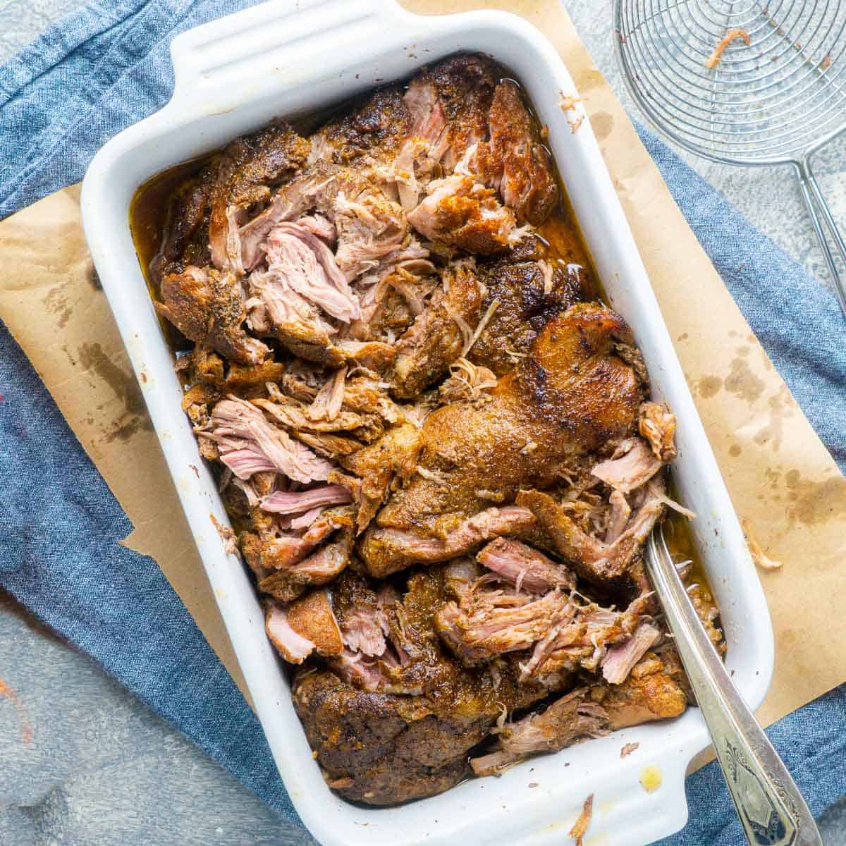 Slow cooker pulled pork with two forks in white serving dish on brown paper next to wire strainer