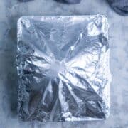 A square baking dish covered with aluminum foil with a blue linen nearby.
