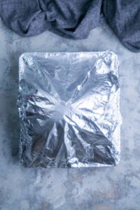 A square baking dish covered with aluminum foil with a blue linen nearby.