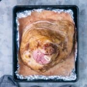 A cooked spiral ham rests in a foil-lined baking tray, with a meat thermometer and bowl of sauce placed nearby on a gray surface.