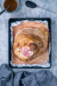 A cooked spiral ham rests in a foil-lined baking tray, with a meat thermometer and bowl of sauce placed nearby on a gray surface.