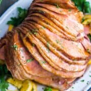 Juicy, glistening, glazed spiral sliced ham served on a white platter, garnished with parsley and orange slices.