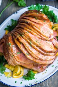 Juicy, glistening, glazed spiral sliced ham served on a white platter, garnished with parsley and orange slices.