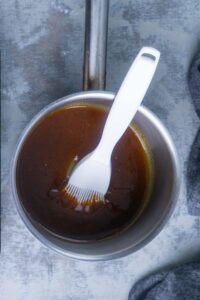 A saucepan filled with ham glaze and a white silicone basting brush resting inside on a gray surface.