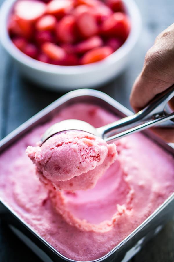 Strawberry frozen yogurt being scooped from a stainless silver container with an ice cream scoop and white bowl of frozen strawberries on gray wood background