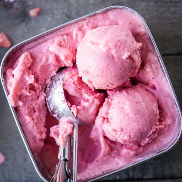 Frozen strawberry yogurt in stainless square container with 3 scoops yogurt and one scoop inside ice cream scoop on gray wood background