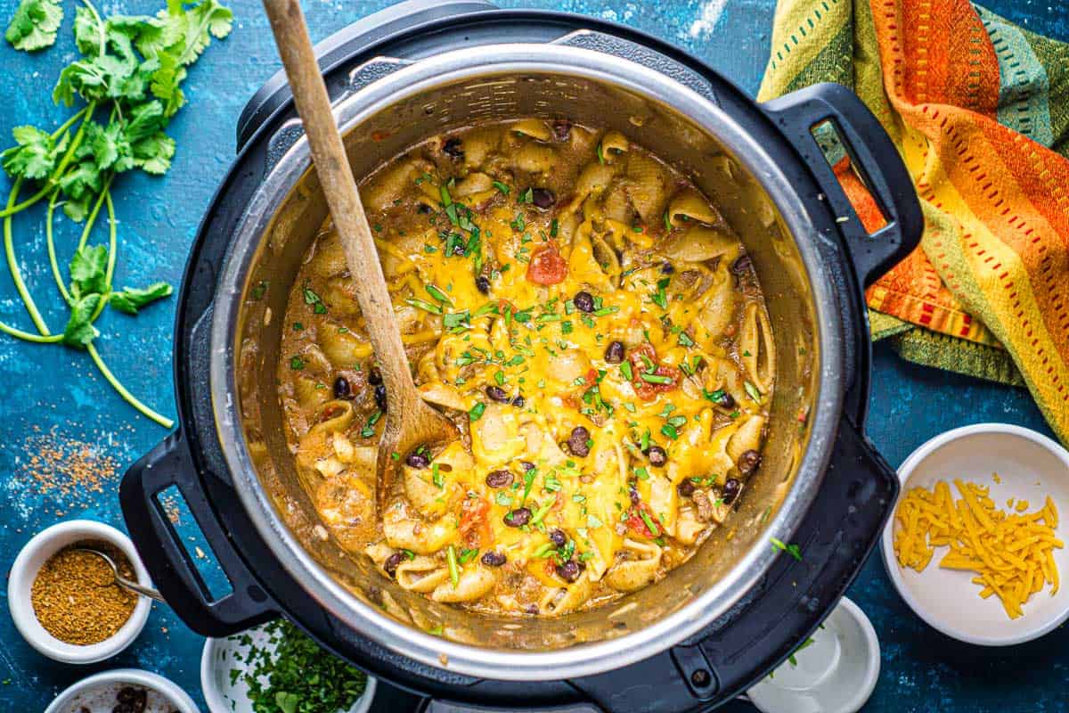 cheese-topped taco pasta in the Instant Pot on blue surface next to small bowls of garnish and colorful napkin