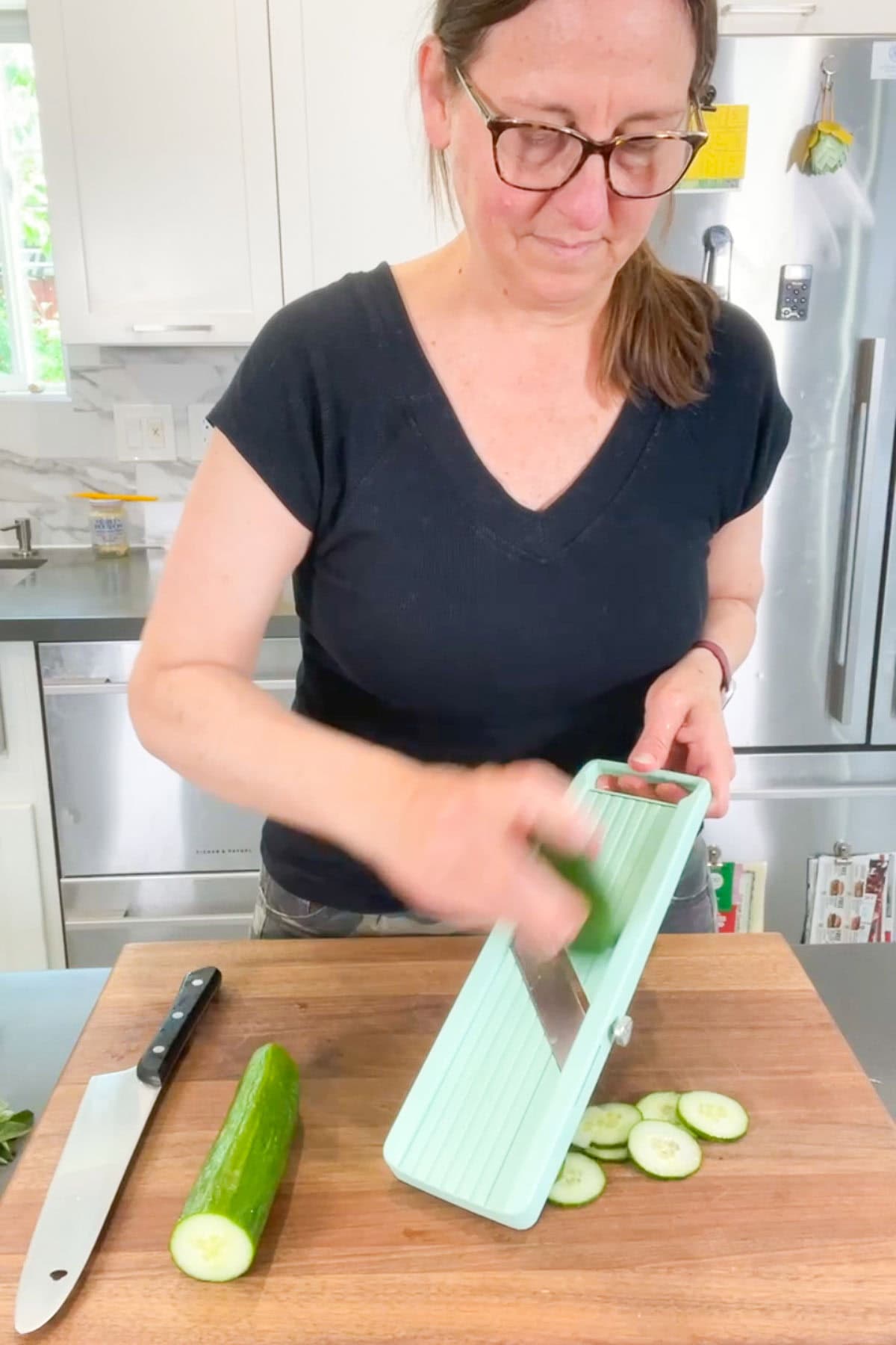 A woman in glasses slices a cucumber using a mint green mandoline on a wooden cutting board in a kitchen.