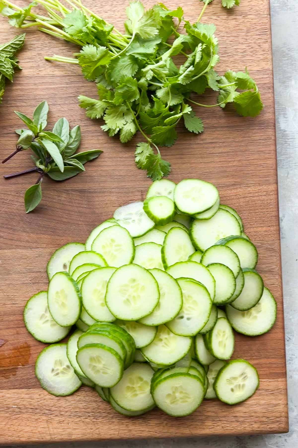 Thinly sliced cucumbers are arranged on a wooden cutting board next to sprigs of fresh cilantro and Thai basil.
