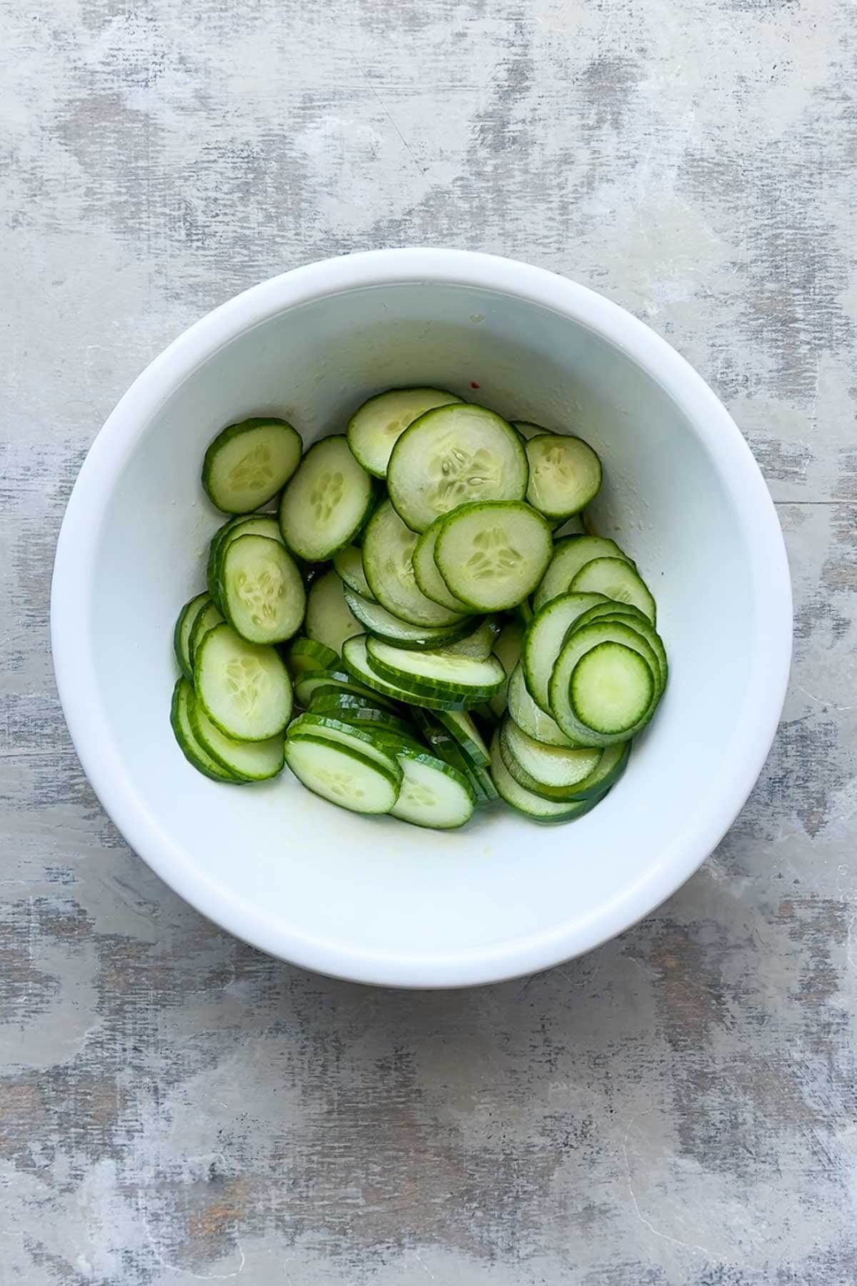 A white bowl filled with thinly sliced cucumbers on a light gray textured surface.