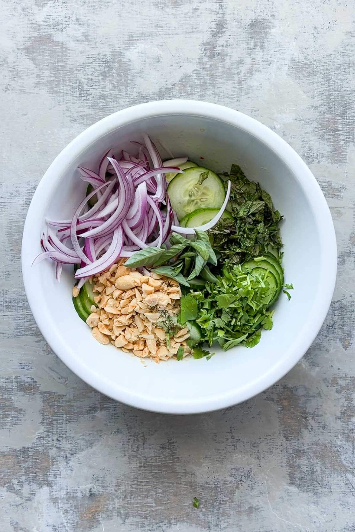 A white bowl contains sliced red onions, cucumber, fresh herbs, chopped peanuts, and green chili peppers, arranged in separate sections.
