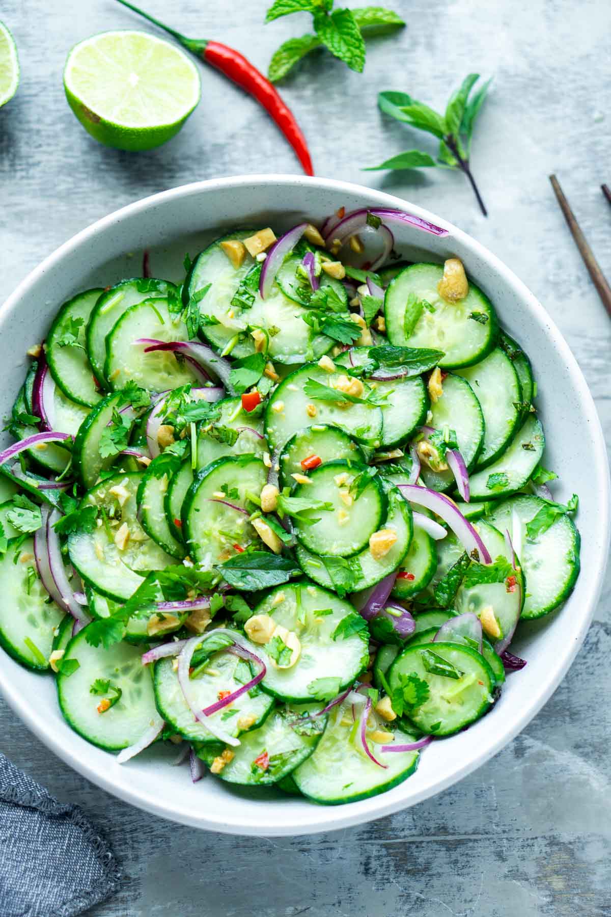 A bowl of Thai cucumber salad with red onion, herbs, chopped nuts, and Thai chili, placed on a light surface with a pair of chopsticks nearby.