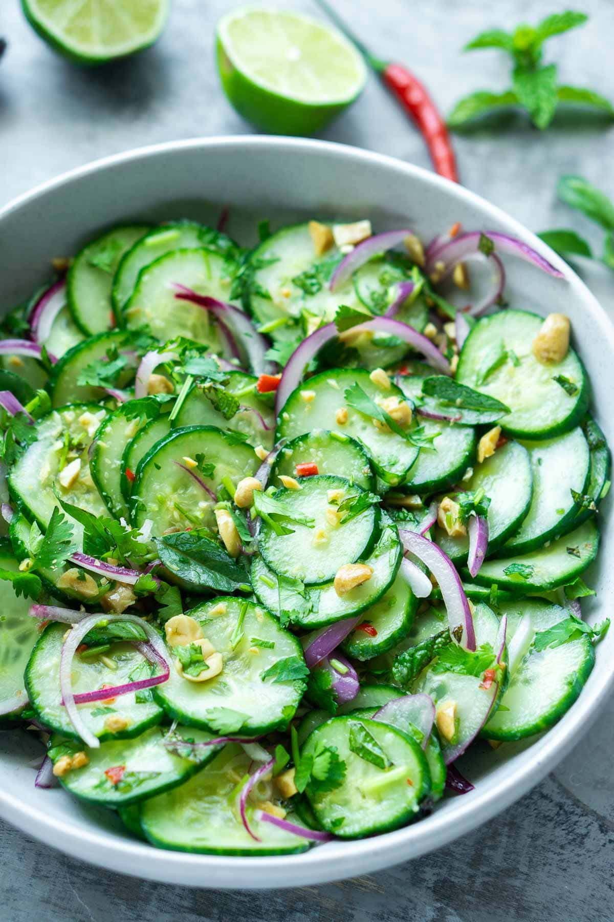 A bowl of Thai cucumber salad with red onion, herbs, chopped nuts, and Thai chili, placed on a light surface.