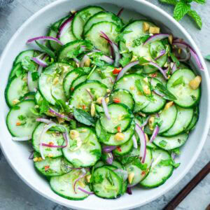 A bowl of Thai cucumber salad with red onion, herbs, chopped nuts, and Thai chili, placed on a light surface.