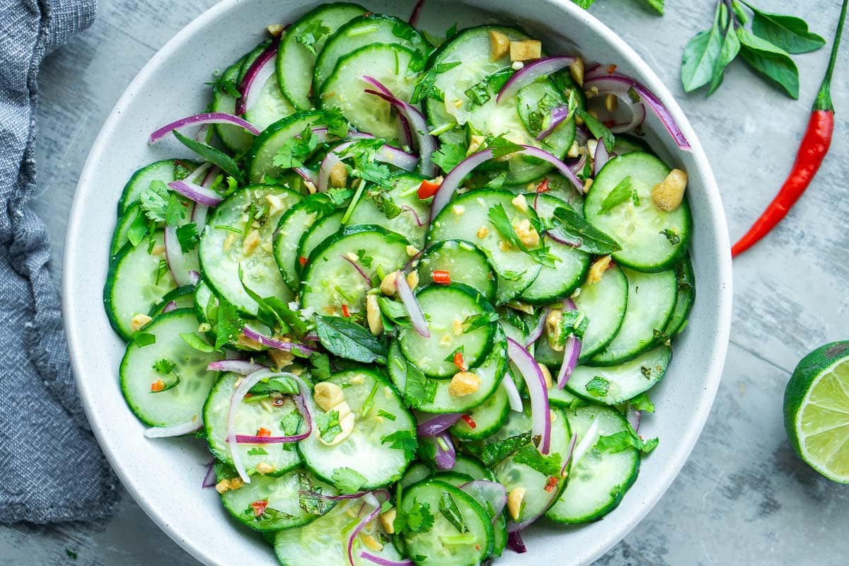 A bowl of Thai cucumber salad with red onion, herbs, chopped nuts, and Thai chili, placed on a light surface.