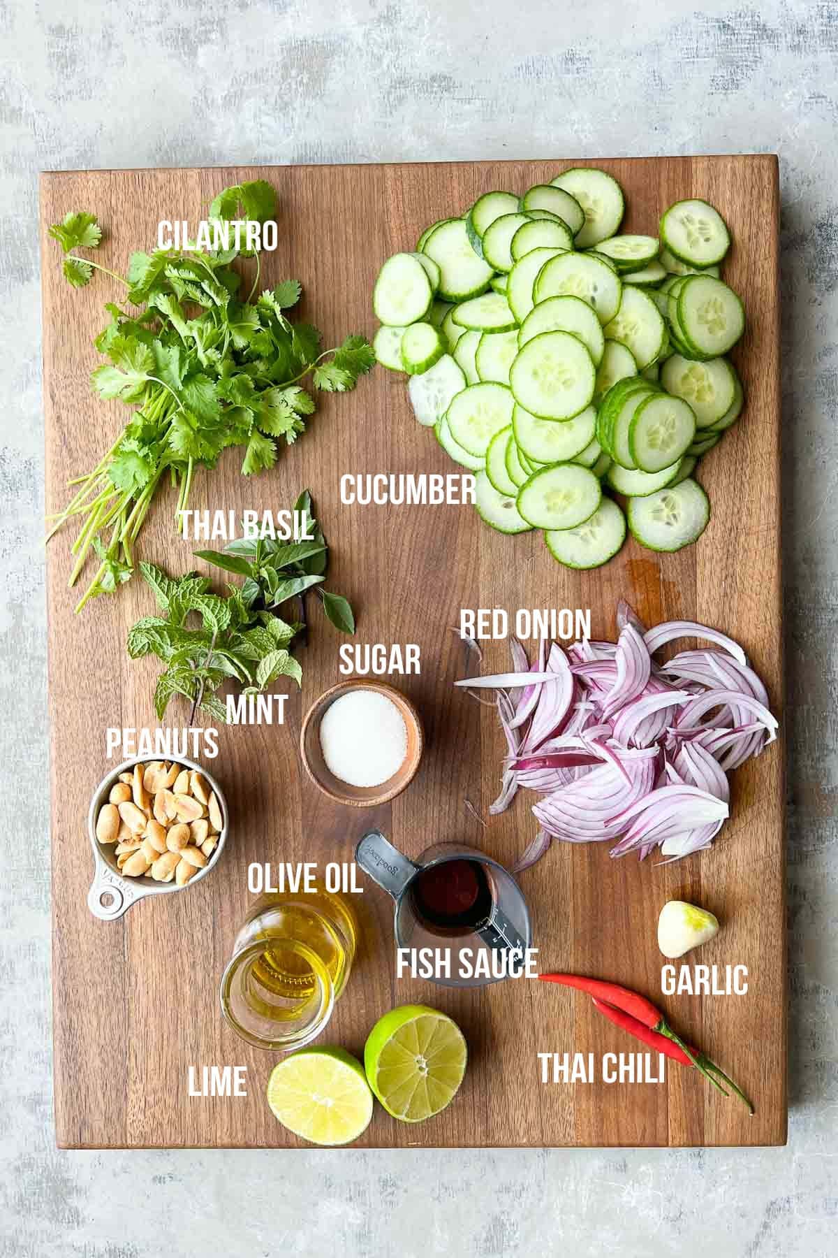 Wooden board with labeled ingredients: cilantro, basil, mint, cucumber, red onion, peanuts, lime, olive oil, fish sauce, sugar, garlic, and Thai chili.