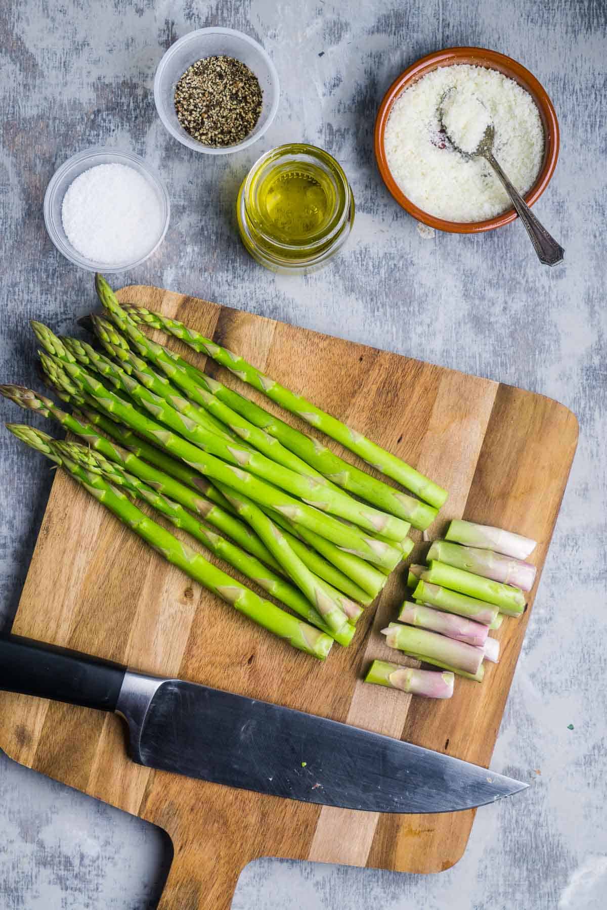trimmed asparagus and chef knife on cutting board next to containers of Parmesan, salt, pepper, and oil