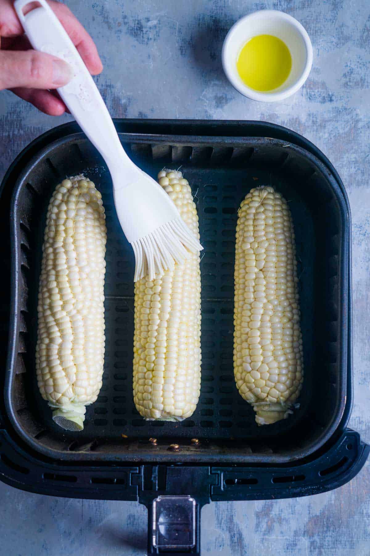 corn on the cob in air fryer basket is brushed with oil