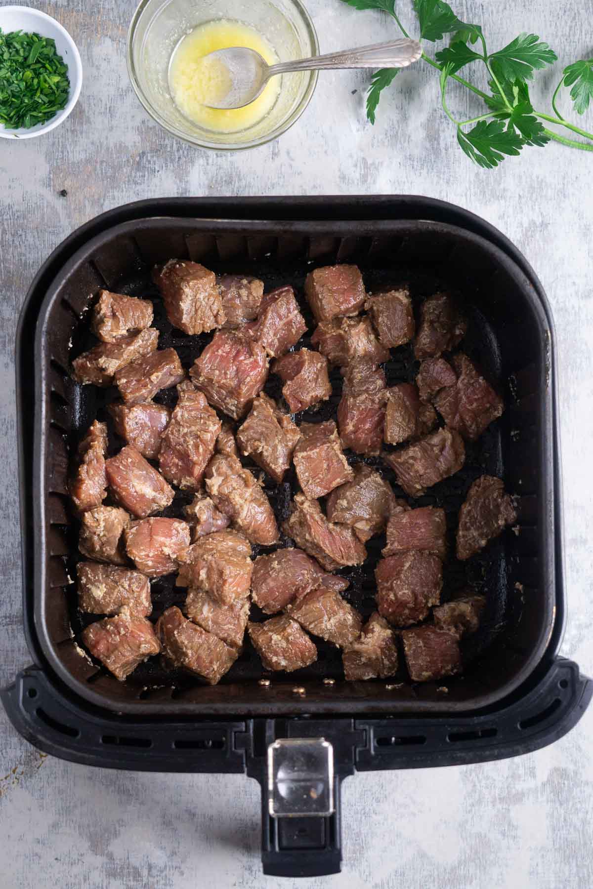 coated steak cubes in air fryer basket next to garlic butter and parsley.