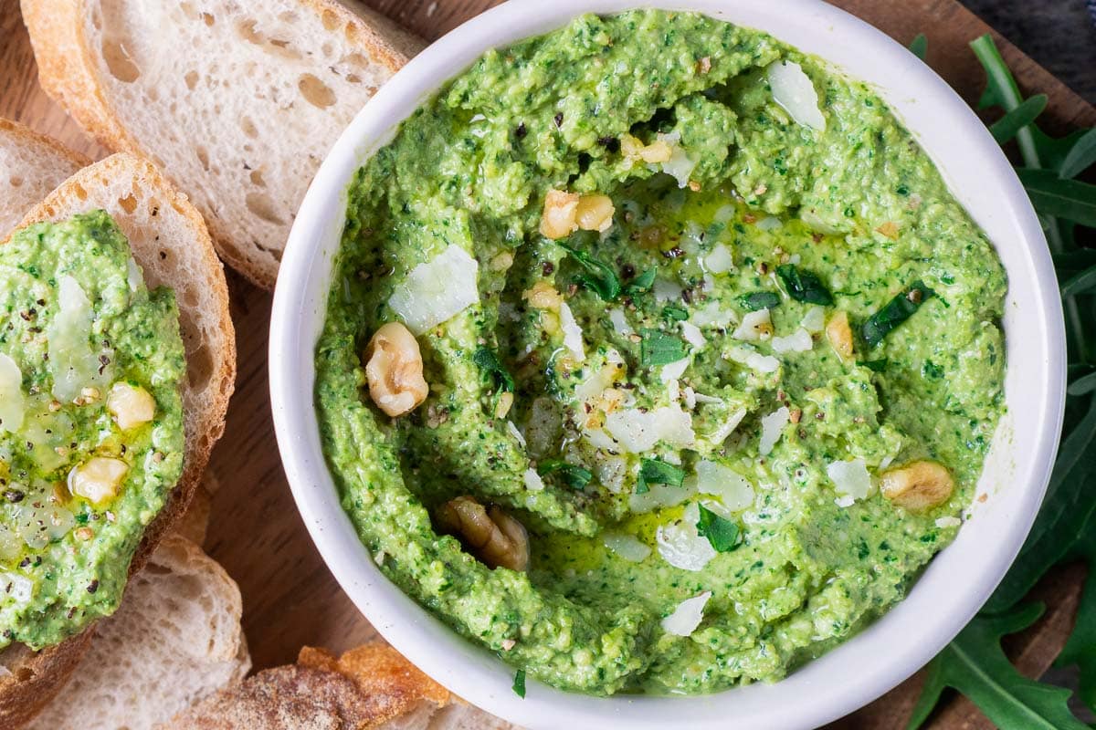 A bowl of arugula pesto sauce topped with walnuts, grated cheese, herbs, and olive oil, served with slices of crusty bread.