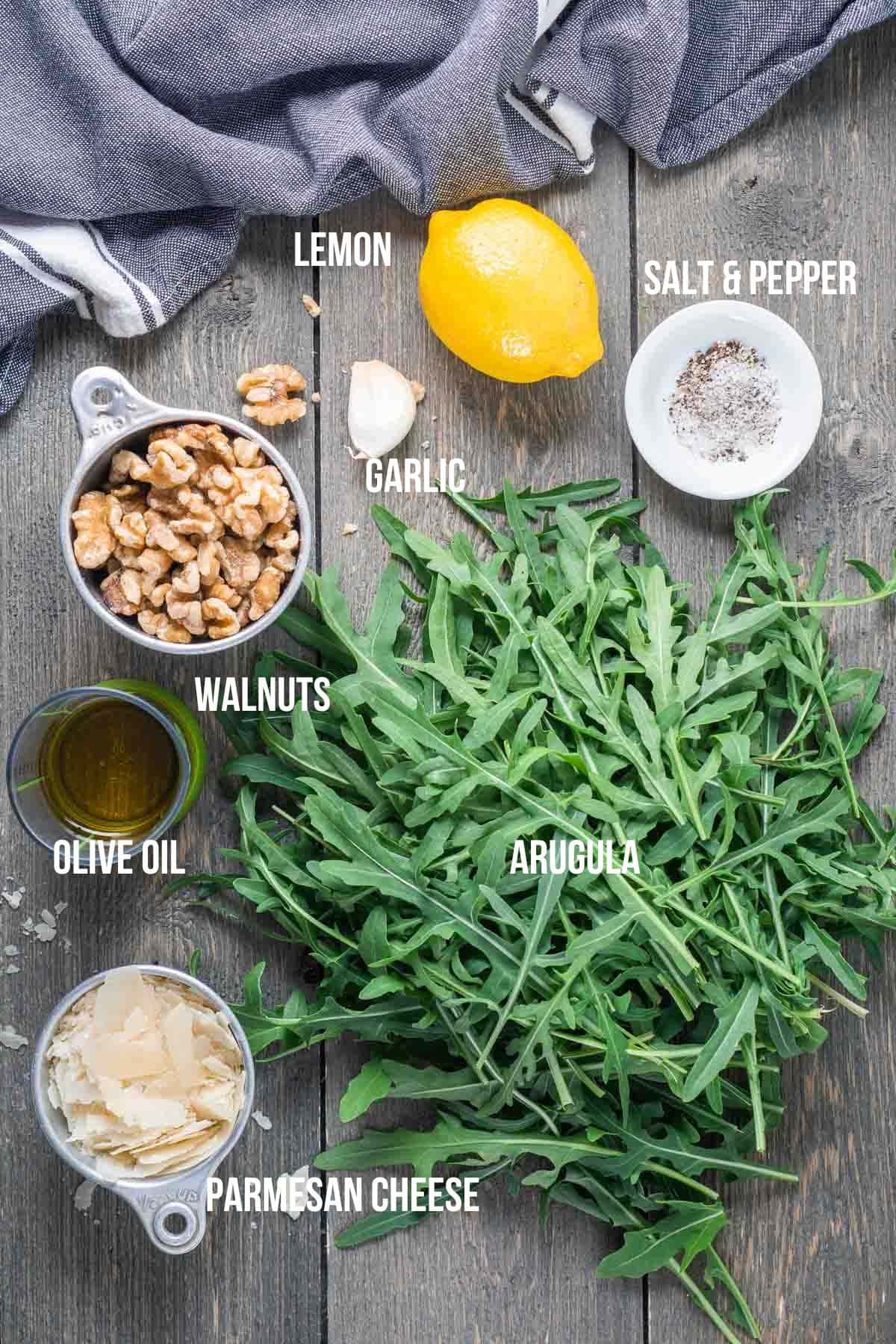 Ingredients for arugula pesto are arranged on a wooden surface: fresh arugula, walnuts, parmesan cheese, olive oil, garlic, lemon, and a small bowl of salt and pepper.
