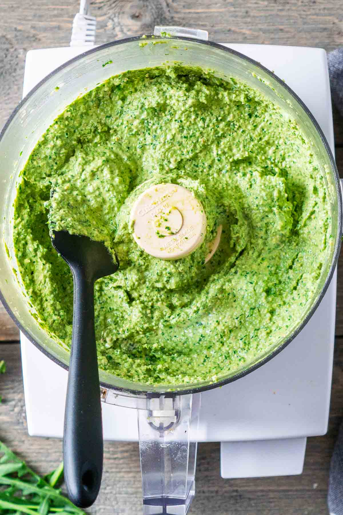 A food processor filled with arugula pesto with a black spatula resting inside.
