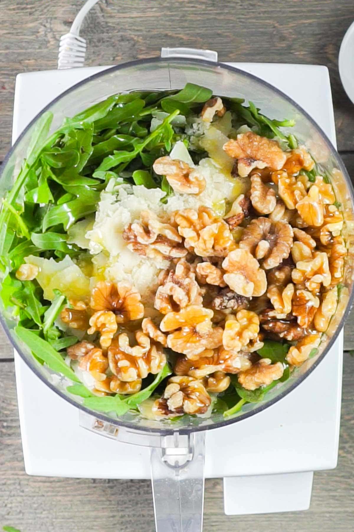 A food processor filled with arugula, walnuts, grated cheese, garlic, and olive oil on a wooden surface.