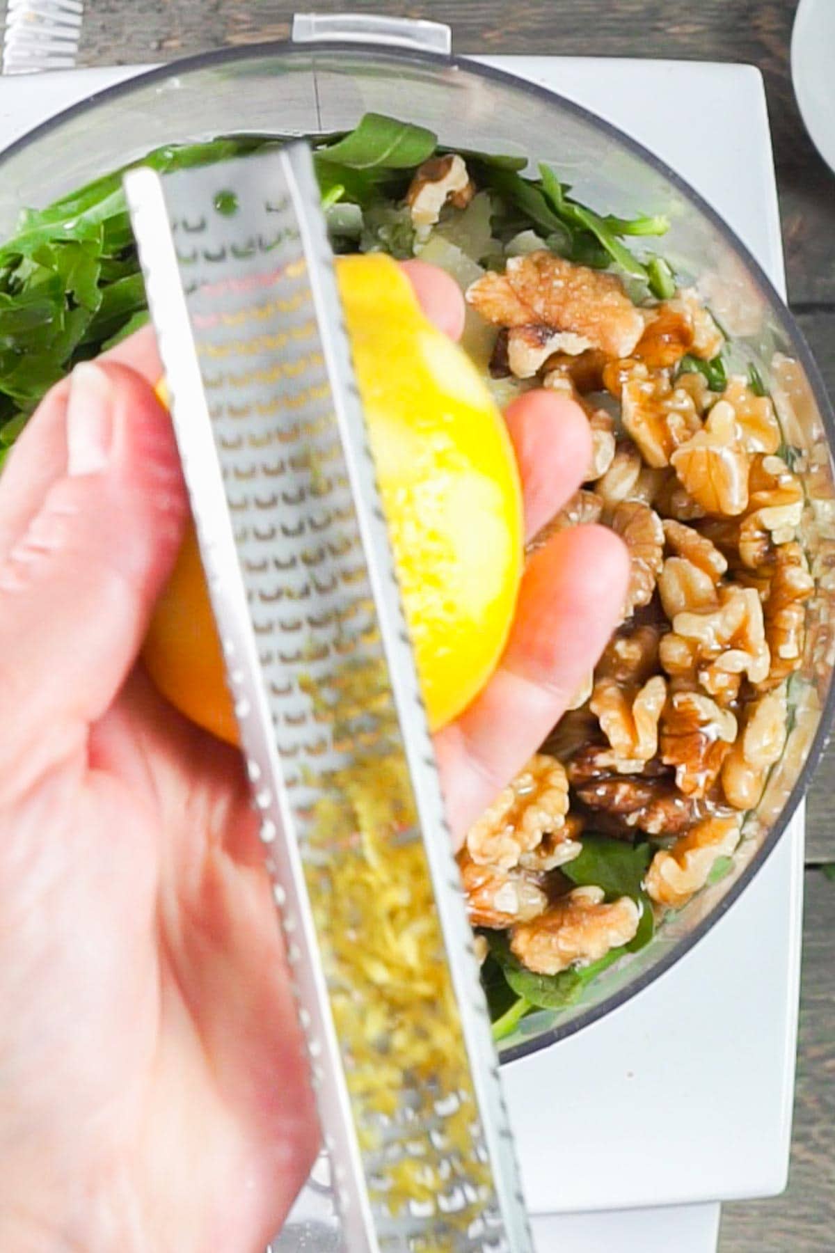 A hand zesting a lemon over a food processor bowl containing walnuts and arugula leaves.
