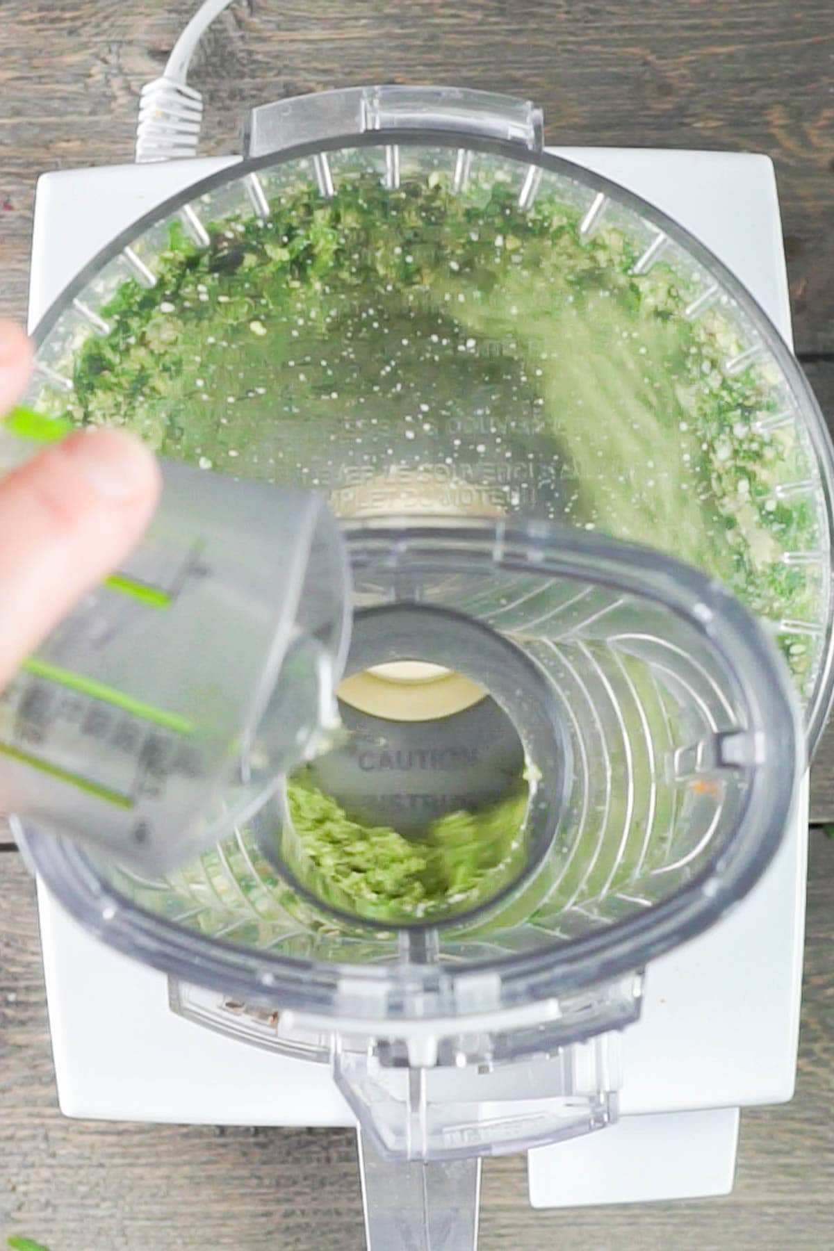 A hand pours liquid from a measuring cup into a food processor blending green ingredients for fresh arugula pesto.