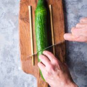 cucumber between two wooden chopsticks on cutting board is being sliced by knife in a person's hands