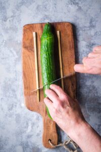 cucumber between two wooden chopsticks on cutting board is being sliced by knife in a person's hands