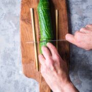 cucumber between two wooden chopsticks on cutting board is being sliced by knife in a person's hands