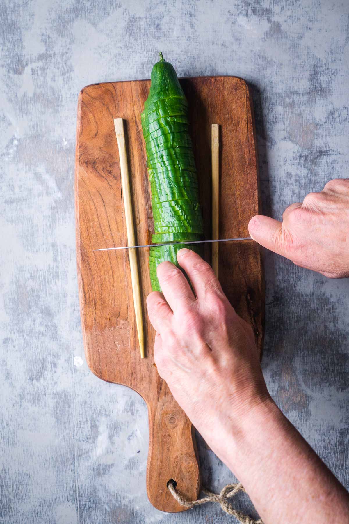 cucumber between two wooden chopsticks on cutting board is being sliced by knife in a person's hands