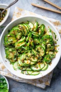 Asian cucumber salad in white bowl on printed napkin next to garnish ingredients
