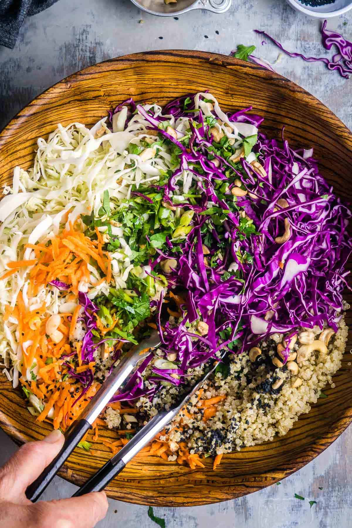 A large wooden bowl filled with Asian slaw&mdash;shredded green and red cabbage, carrots, herbs, quinoa, cashews, and seeds&mdash;with a hand holding tongs mixing the fresh ingredients.