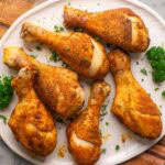 Six seasoned, crispy baked chicken legs are served on a white plate garnished with chopped parsley.