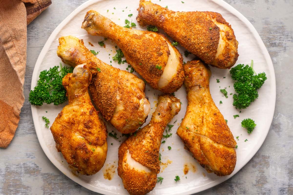Six seasoned, crispy baked chicken legs are served on a white plate garnished with chopped parsley.