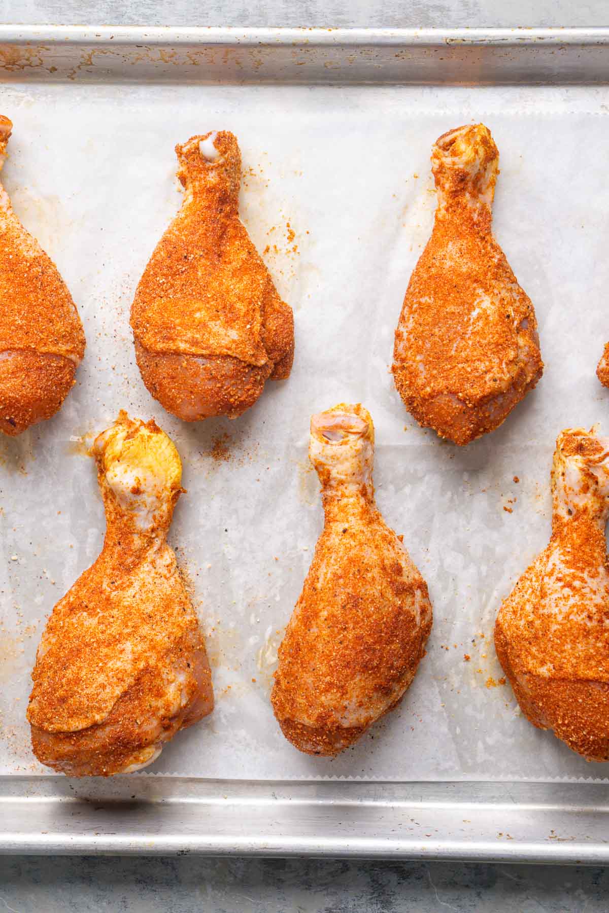Raw chicken leg drumsticks coated with seasoning are arranged on a parchment-lined baking sheet.