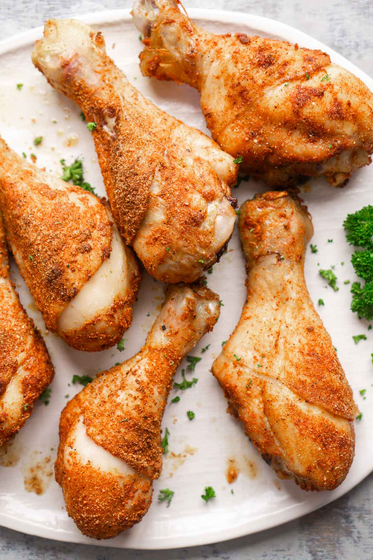 Six crispy baked chicken legs are served on a white plate garnished with chopped parsley.