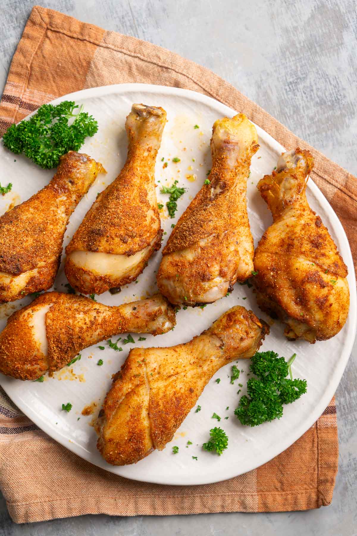 Six crispy baked chicken legs are served on a white plate garnished with chopped parsley.