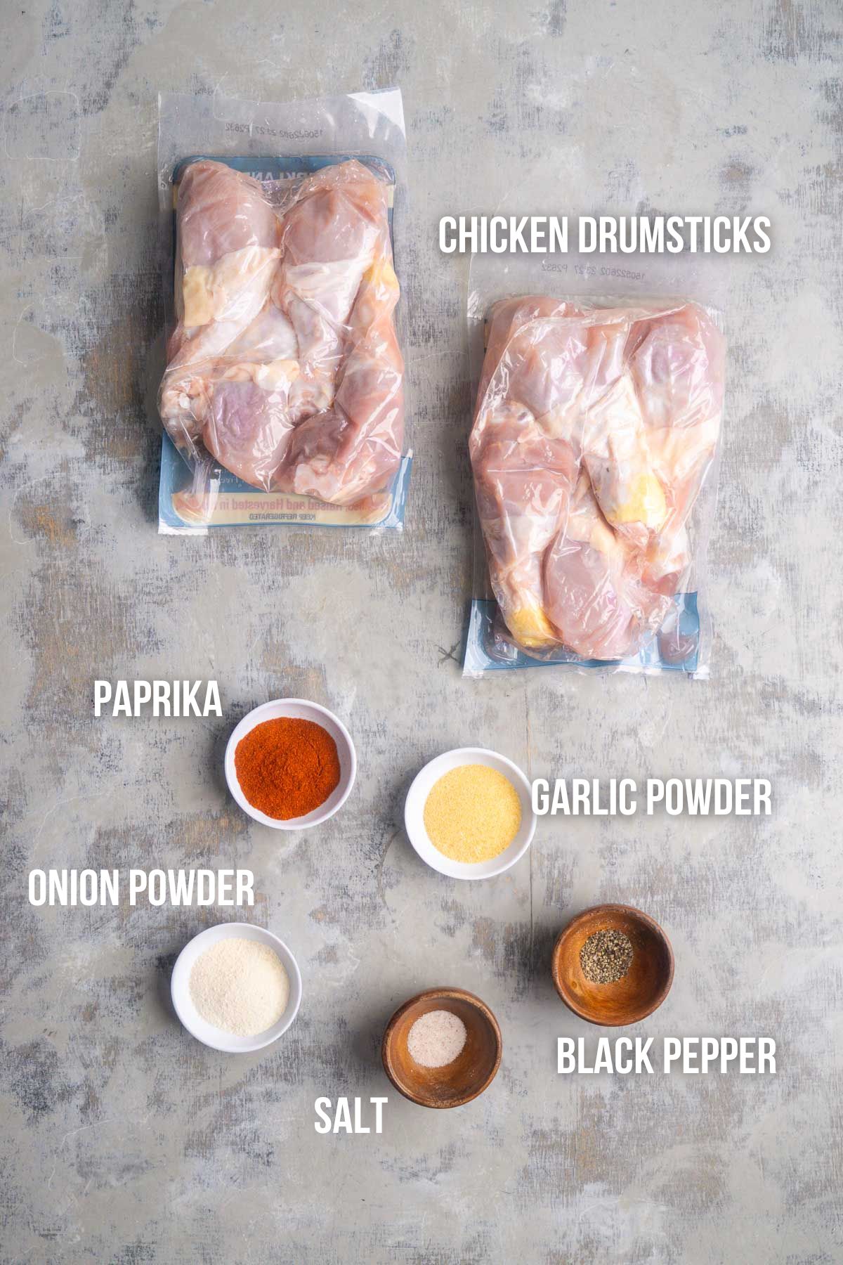 Two vacuum-sealed packs of chicken drumsticks and six small bowls containing paprika, garlic powder, onion powder, salt, and black pepper are arranged on a gray surface&mdash;perfect ingredients for flavorful baked chicken legs.